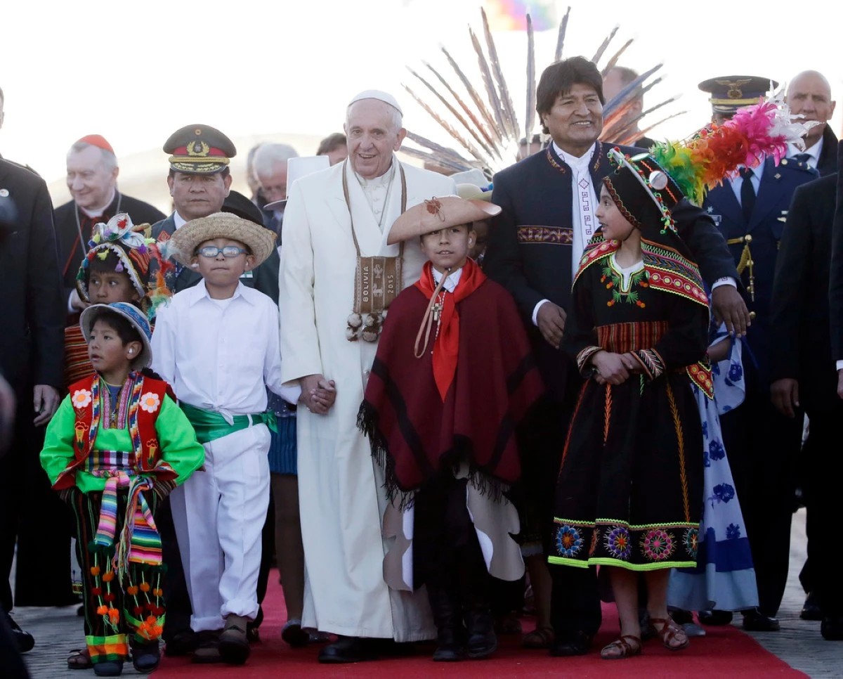 FILE - Pope Francis holds hands with children wearing traditional costumes as he walks with Bolivian President Evo Morales upon his arrival at the airport in El Alto, Bolivia, July 8, 2015. The pouch Francis is wearing around his neck was given to him by Morales. It's woven of alpaca with Indigenous trimmings and traditionally used by people in the Andes to hold coca leaves. (AP Photo/Gregorio Borgia, File)