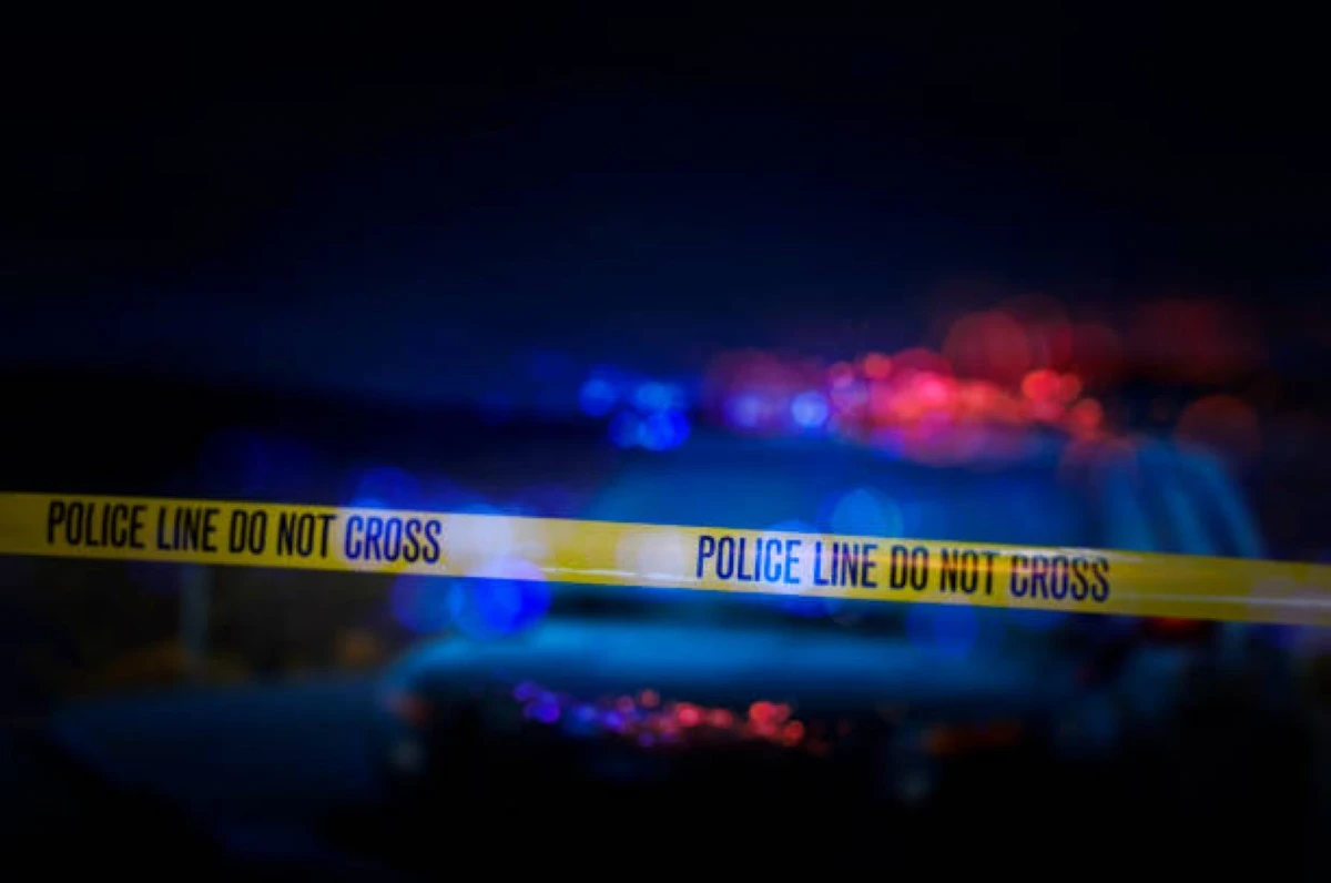 A stock photo of a Police Line "Do Not Cross" caution tape with a defocused police car with sirens flashing red and blue.