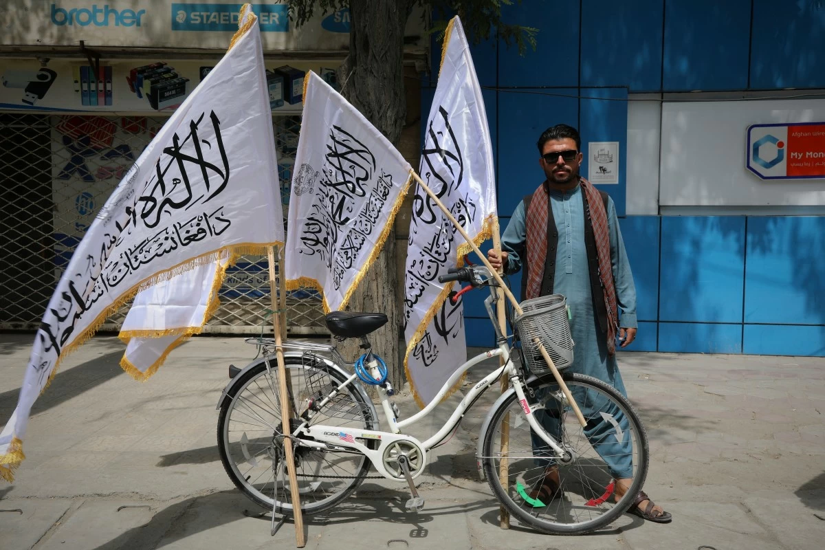 A man poses for the camera next to his bicycle while selling Taliban flags ahead of celebrations marking the fourth anniversary of the U.S. withdrawal from Afghanistan and the beginning of Taliban rule in Kabul, Afghanistan, Thursday, Aug. 14, 2025. (AP Photo/Siddiqullah Alizai)