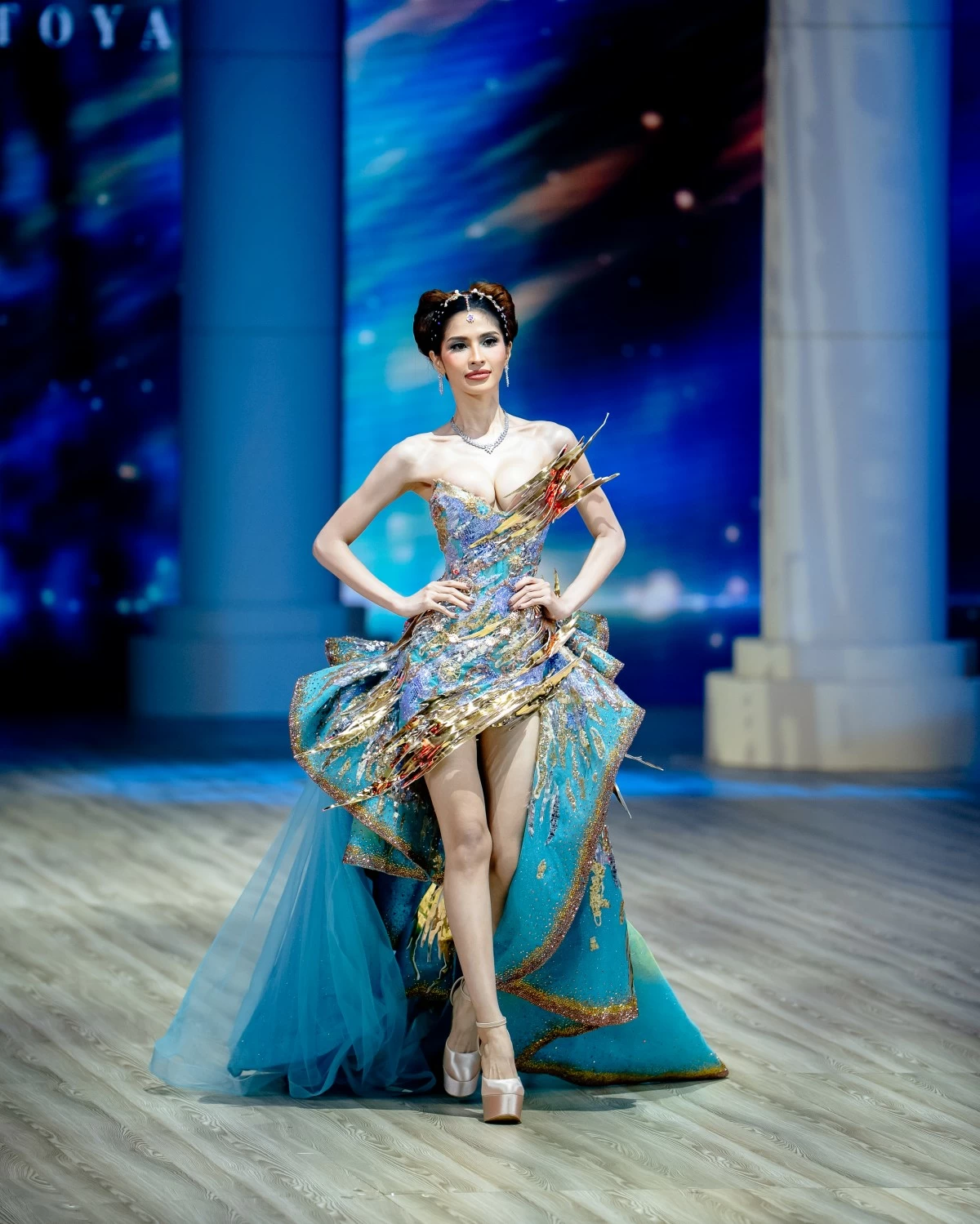 A sculptural short dress with an asymmetric cascading side train is adorned with intricate beadwork in celestial hues, crowned with towering silhouettes and metallic gold overlays. 